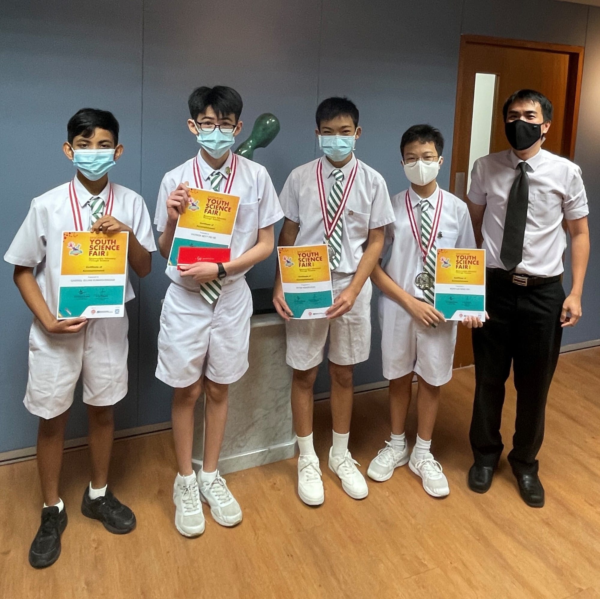 Four masked students in white uniforms hold Youth Science Fair certificates and medals, standing with a masked adult teacher.